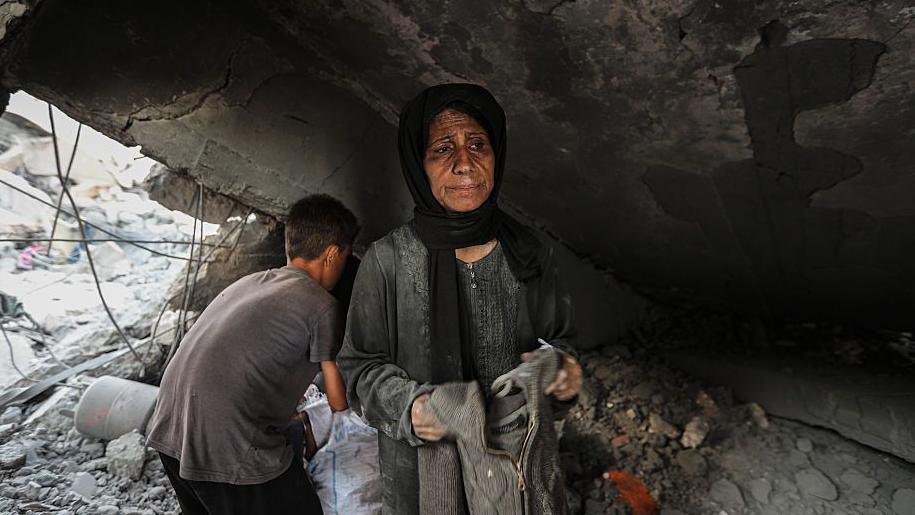 A woman win a dark headscarf and grey dress holds a jumper in her hands as she goes through items in rubble found at a market in Gaza that was struck by Israel. A boy is behind her with his back to the camera.