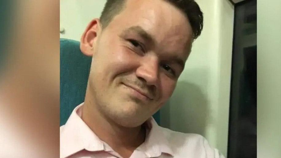 Damien Walton has dark hair, shaved as the sides. He wears a light, collared shirt and stands in a room, smiling at the camera.