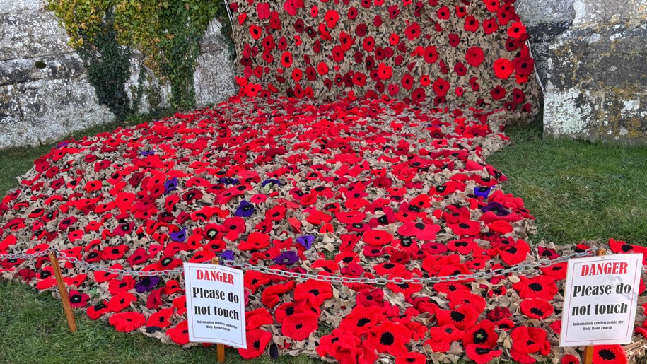 A close-up of the poppy installation at ground level. Hundreds of knitted poppies are attached to the cargo net which is puddling at the bottom of the tower. A small chain has been placed around the artwork with signs saying - danger, please do not touch.