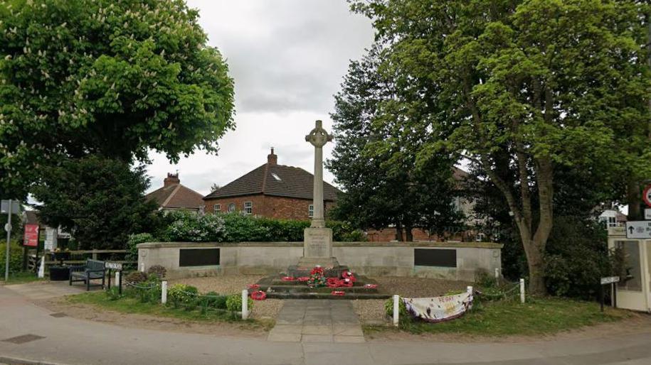 A war memorial in Brough. Poppy wreaths are placed in front of it. Two large trees are at either side of the memorial. The sky is overcast. 