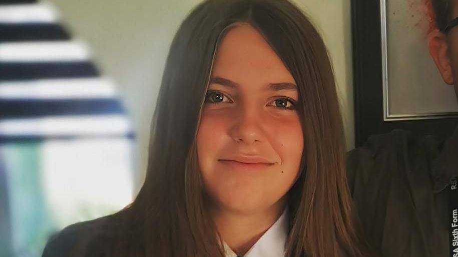 Chloe Longster with long dark brown hair smiling at the camera wearing a white shirt and black jacket. There is a framed picture behind her.