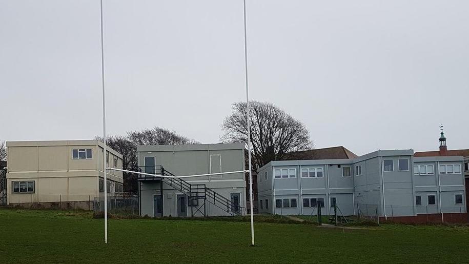 Three temporary modular buildings in the background and a chain-link fence between them and a local park. 