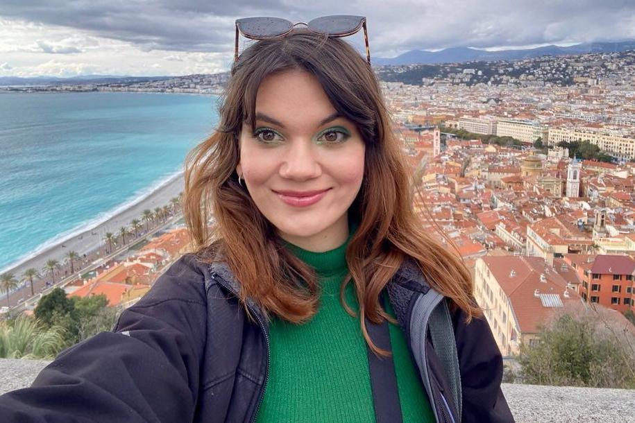 A woman is smiling for a selfie from a tower. These is a city, mountains and the sea in the background