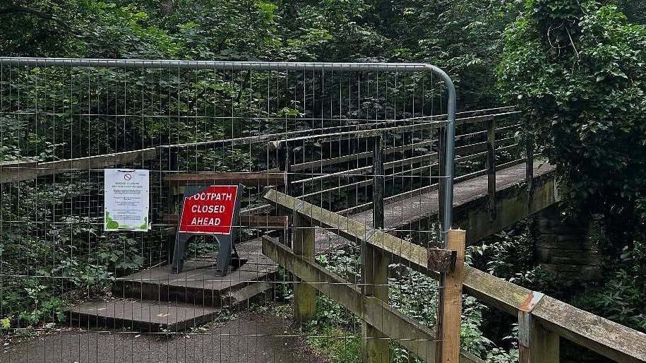 A metal gate is blocking the entrance of the wooden bridge at Cherry Tree Walk in Jesmond Dene. A red sign reading FOOTPATH CLOSED AHEAD in white is place behind the gate, where the narrow wooden footbridge begins. The structure is surrounded by trees and shrubs.