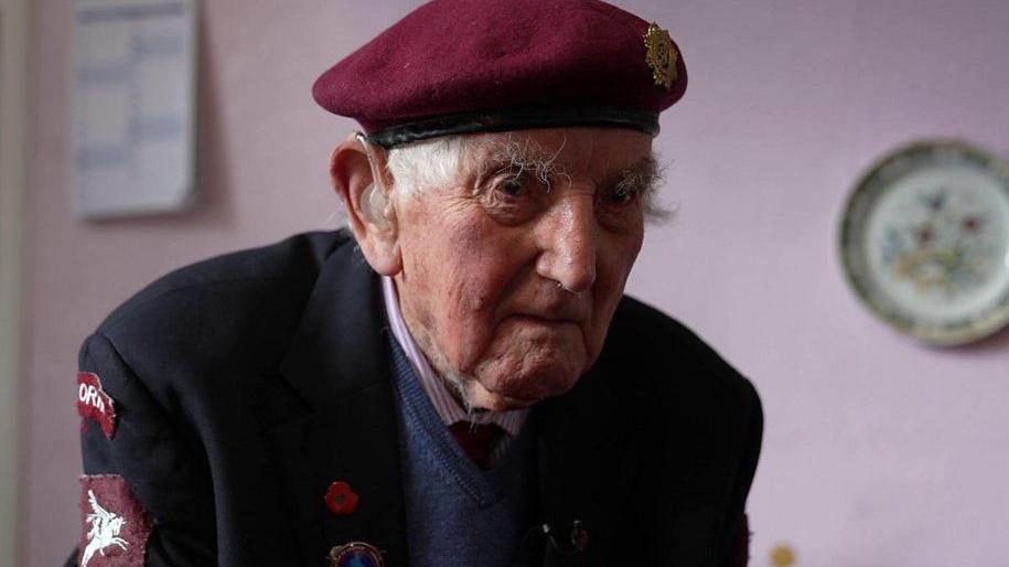 An elderly man wearing a veteran's beret and jacket. The burgundy beret has a gold military badge on it. The jacket has military patches on the sleeve and a red poppy badge on the lapel. Behind the man is a pink wall with a calendar and a plate on it.