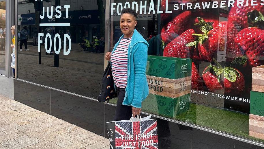 Woman looking into camera while standing outside M&S shop with an M&S bag in her hand.
