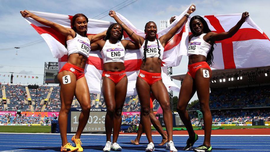 England's Asha Philip, Imani Lansiquot, Biancsa Williams and Daryll Neita stand on the the running track after winning the silver medal in the Women's 4 x 100m Relay. Arm in arm they raise an England flag behind them.