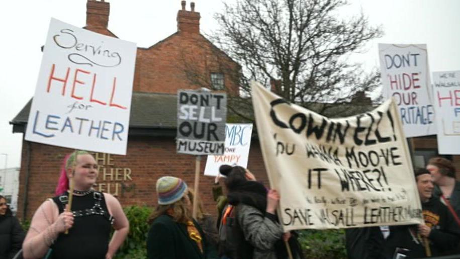 A group of protestors stand outside the Walsall Leather Museum, a redbrick building with a tree and bushes outside. The protestors carry various campaign signs and banners, including 'Don't Sell Our Museum' and 'Serving Hell for Leather'