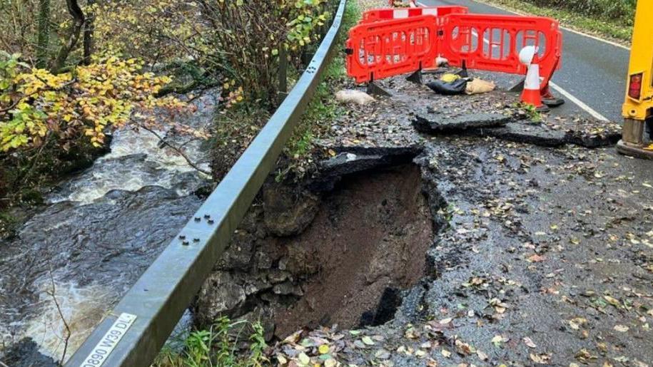 A hole in the road with red cones surrounding it. Below, a nearby river flows past. 