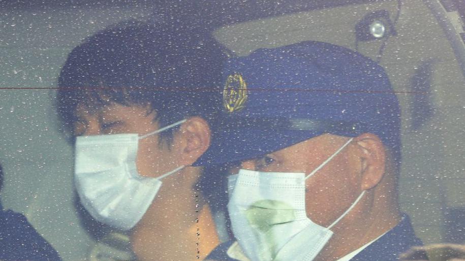 A young man and a police officer in a blue cap, both wearing face masks, seen through a window.