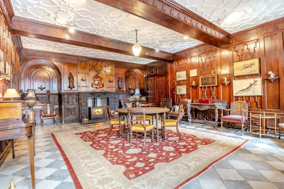 Wood panelling including what looks like a heraldic coat of arms and two carved images of men can be seen in a large room with a carved wooden fireplace. The room includes furniture such as wooden tables and chairs.