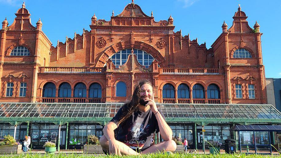 Matt Panesh sitting cross-legged on grass outside the Winter Gardens in Morecambe