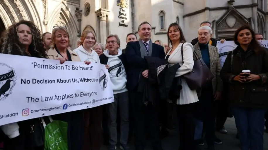 A group of people stand outside the Royal Courts of Justice holding a banner that reads "permission to be heard - no decision about us without us - the law applies to everyone".