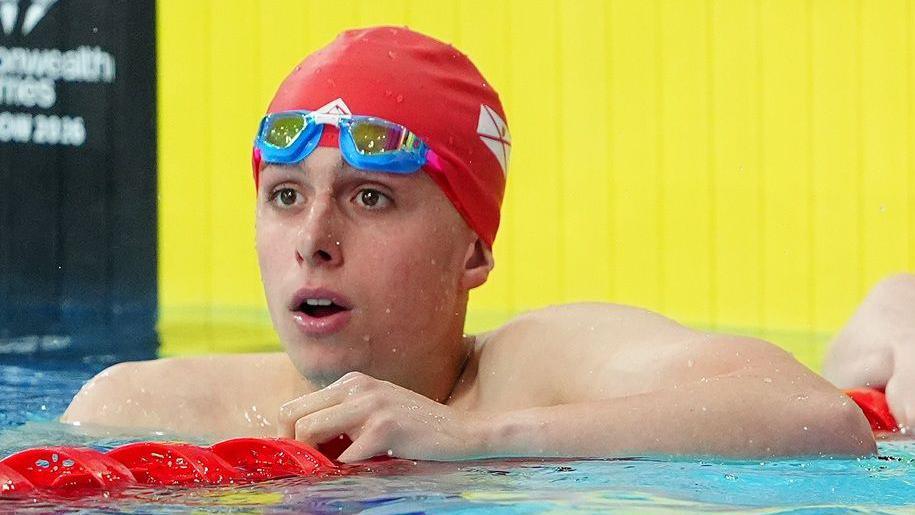 Filip Nowacki, wearing a swimming hat and raised goggles, waits in the pool for the result of a race