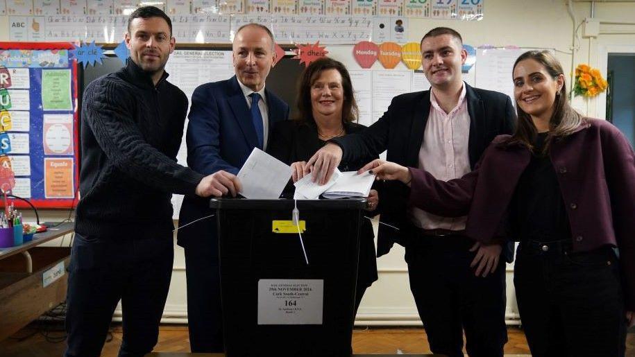 Micheál Martin stands with his family in a primary school classroom as the members place their white ballot papers into a black vote box. 