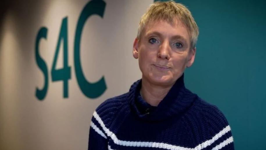 Sian Doyle poses against wall which is painted white and blue and has S4C written on it. She wears a navy jumper with white stripes and has short, blonde hair.