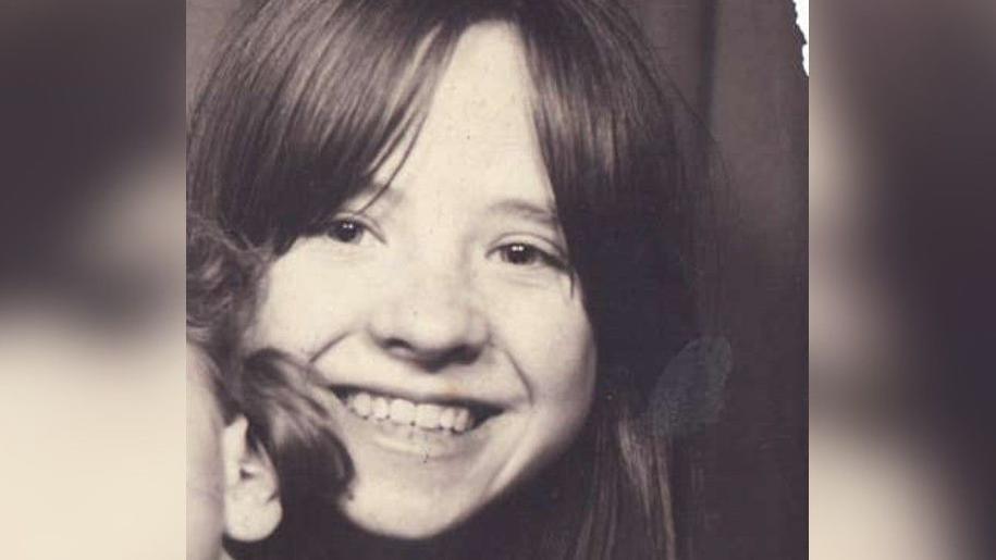 Kathleen as a girl, in a black and white image. She is smiling at the camera and has brown hair, with a fringe that is parted in the middle. She has a child on her lap but they are out of shot, with just an ear and a bit of hair showing.