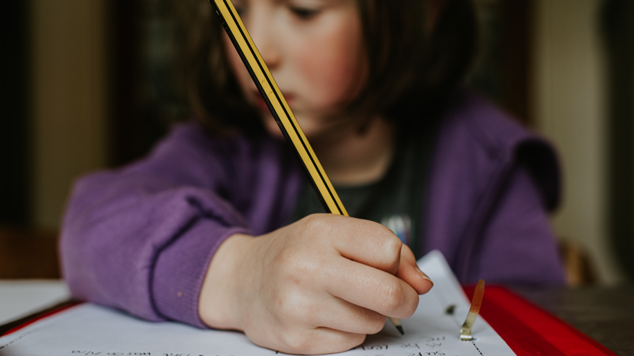 A young girl studying. Her face is blurred but she is holding a pencil over a piece of paper.