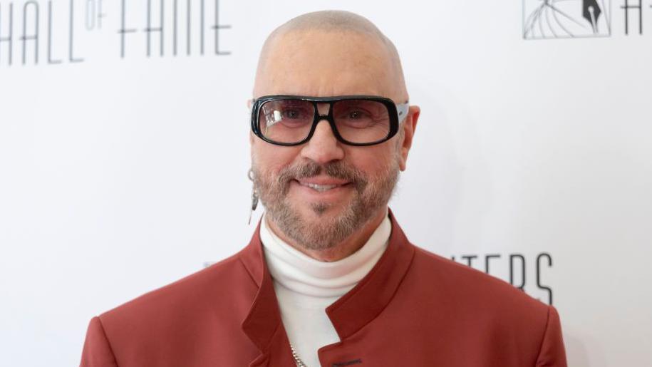A bald man with grey facial hair wearing one earring and sunglasses, as well as a white polo kneck jumper and red jacket.
