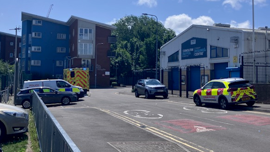 A road with a block of flats at one end and cars parked either side, including a fire service car, a police van and an ambulance with its blue lights on.