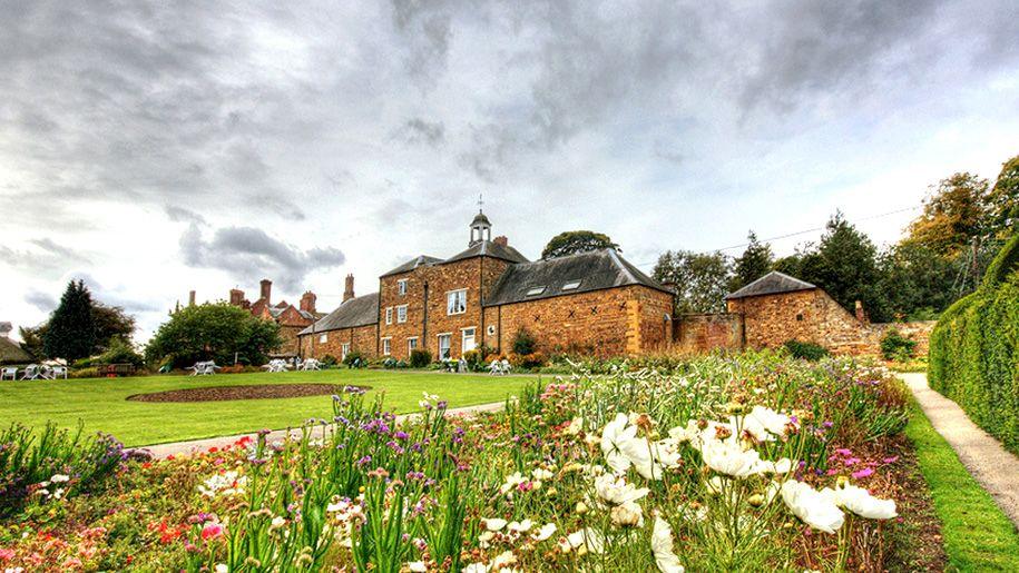 Delapre Abbey, a large brick-built house, with a grey roof, a large garden, with flower beds, and green lawns. 