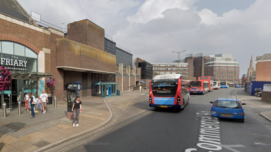 The entrance to Guildford bus station with the Friary Centre pictured on the left. A number of buses can be seen flowing into the bus station, alongside a blue car. A number of people with faces blurred out can be seen near the Friary Centre.