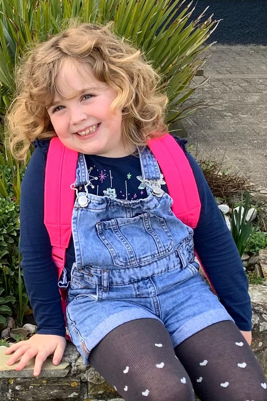Amber is sitting on a low stone wall with plants behind her. She has blonde curly hair and blue eyes. She is wearing denim dungaree shorts, a bright pink backpack and black tights with white hearts.