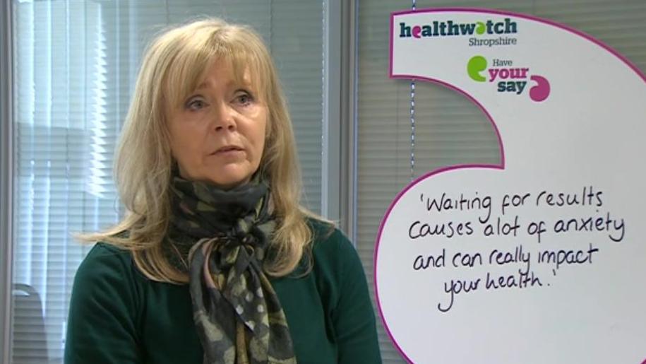 Lynn is sat looking towards the camera - she's wearing a dark green top and a patterned green scarf and has blonde hair past her shoulders. To her left is a Healthwatch Shropshire-branded sign that reads "waiting for results causes a lot of anxiety and can really impact your health".