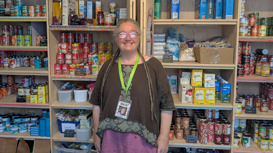 Kit Stevens smiling at the camera. She is wearing a brown top with pink trousers. She is standing in front of shelves filled with different items such as tinned food and tea bags. She is also wearing a lime green lanyard.