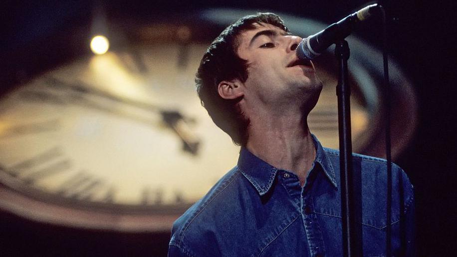 Liam Gallagher, with short hair and a blue denim shirt, sings into a microphone in front of a blurred image of a clock face