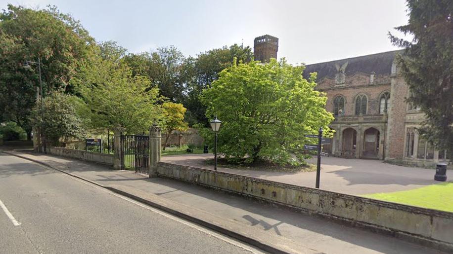 A stone-built, large hall sited next to a main road. Around the hall is a low wall. Trees and shrubs are in the background.