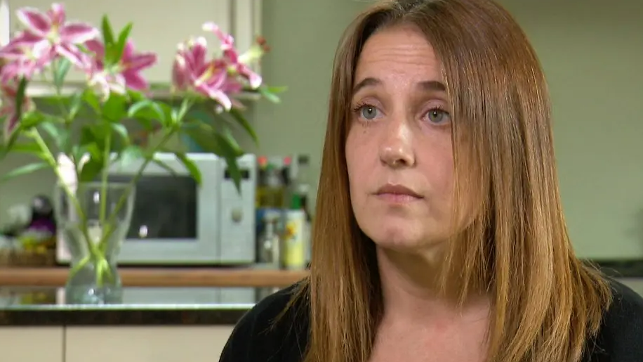 Louise Slorance looks slightly off camera. She has shoulder length light brown hair and is wearing a black cardigan. She is sitting in front of a green wall in her kitchen. There is a vase of pink flowers on the island, which has a black worktop and white cupboards. In the background, there is a silver microwave underneath one cupboard on the worktop, next to a collection of bottles of oil.