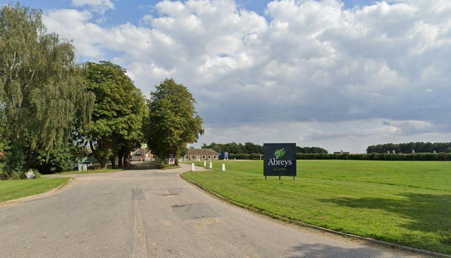 An Abreys sign on the edge of a field of mown grass with a driveway heading past trees towards buildings in the background