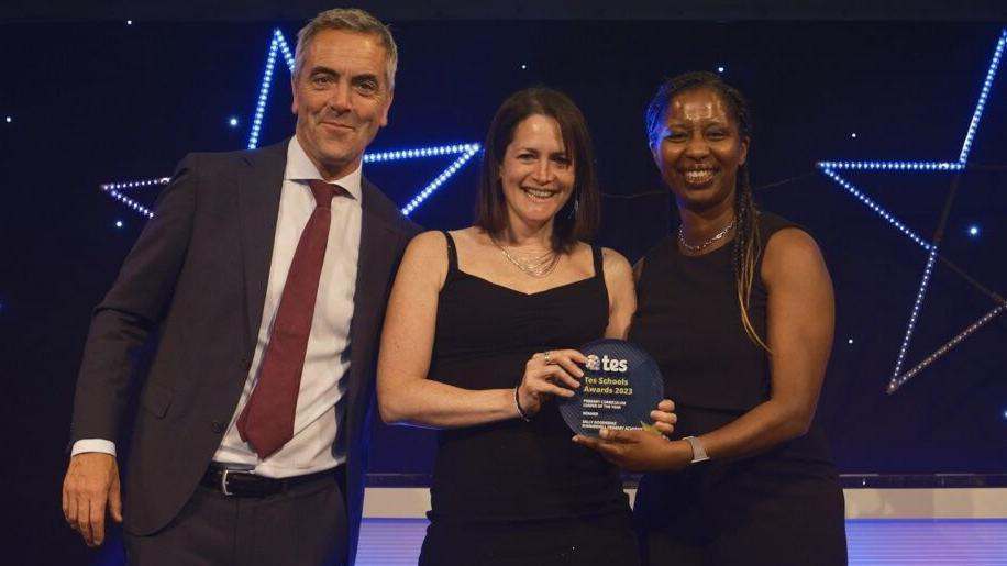 Sally Goodridge wearing a black dress and holding her TES award. She is joined on stage by chief judge Jon Severs on her right, wearing a dark suit and red tie. Another woman is on her left, jointly holding the award and smiling.