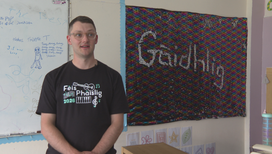 Duine le speuclairean air na sheasamh air beulaibh bratach air a bheil "Gàidhlig" sgrìobhte.
