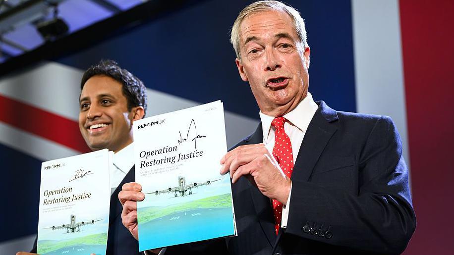 Both are smiling and holding up signed copies of their new policy. The document pictures a plane coming to land on a runway, and reads Operation Restoring Justice. Behind them is the top left corner of a large union jack, that is the backdrop to where they are standing.