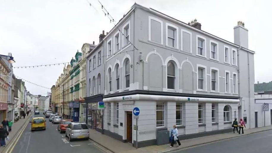 A grey, Victorian-style building on a street corner in Ramsey. Cars can be seen driving along and parked on Parliament Street.