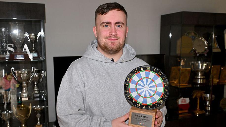 Luke Littler with brown hair and beard wearing a grey hoodie holding a trophy in the shape of a darts board with trophy cabinets either side of him filled with trophies 