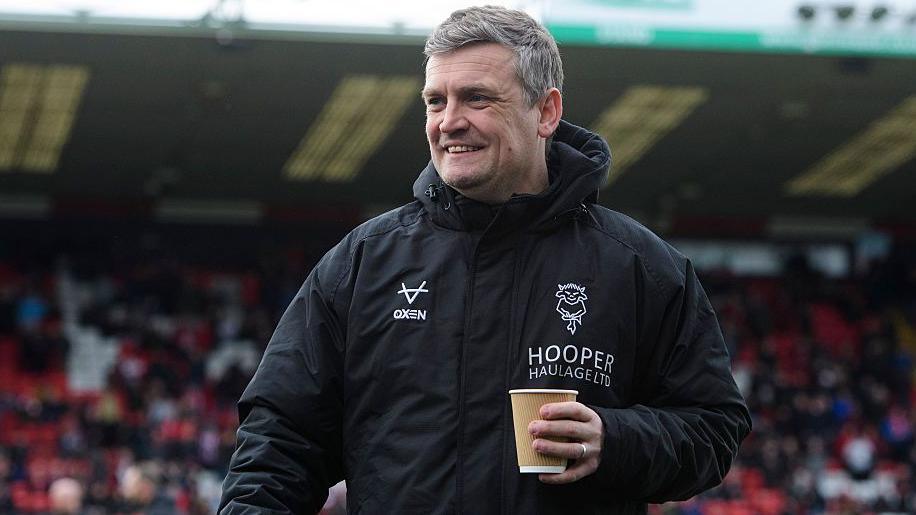 A smiling Lincoln City head coach Michael Skubala wearing a black waterproof Lincoln training top with the club image on it and carrying a cup of coffee in his left hand