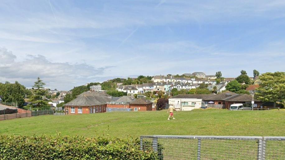 Land for homes off Greenswood Road, Brixham. There is a bush in the front left of the frame and attached to it is a gate. There is a person walking across a field. There are houses in the background. The sky is blue with white clouds.