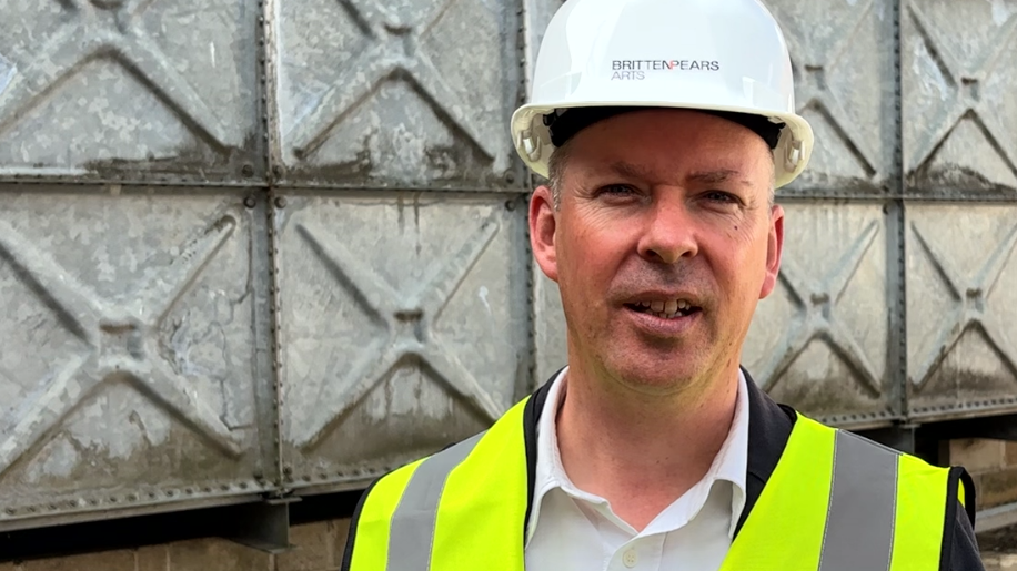 A head and shoulders image of Andrew Comben. He is wearing a white hard hat and a high-vis vest over a white shirt. He is looking into the camera.