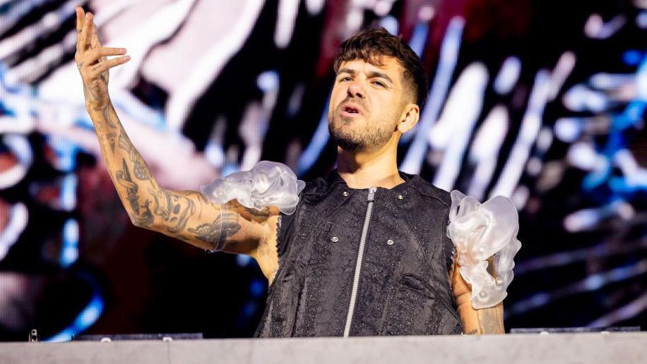 Anyma, with tattooed arms and short dark hair, holds his hand aloft while wearing a black sleeveless top and plastic white shoulder decorations