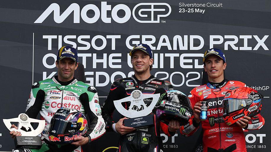 Marco Bezzecchi (centre, wearing black) holds a silver trophy in his right hand and helmet in his left with Johann Zarco (wearing green and white) and Marc Marquez (in red) to his left and right on the podium