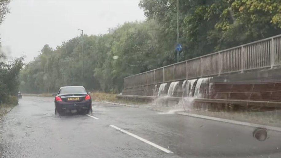 Water flowing under the railings of a raised footway onto the main carriageway, photographed from a car window. Another car appears to be proceeding cautiously through flood water in front, along the middle of the road.