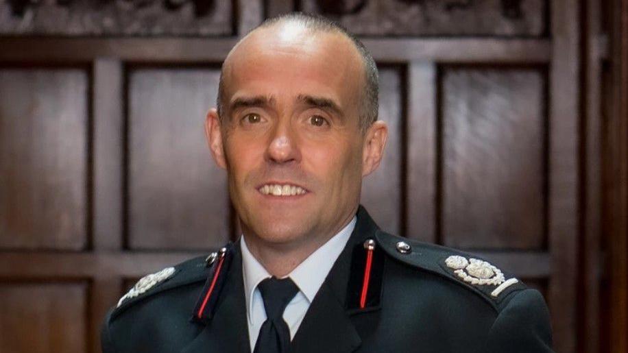 Rob Barber, chief fire officer for Staffordshire Fire and Rescue Service, is stood  against a wood-panelled wall in his uniform. 