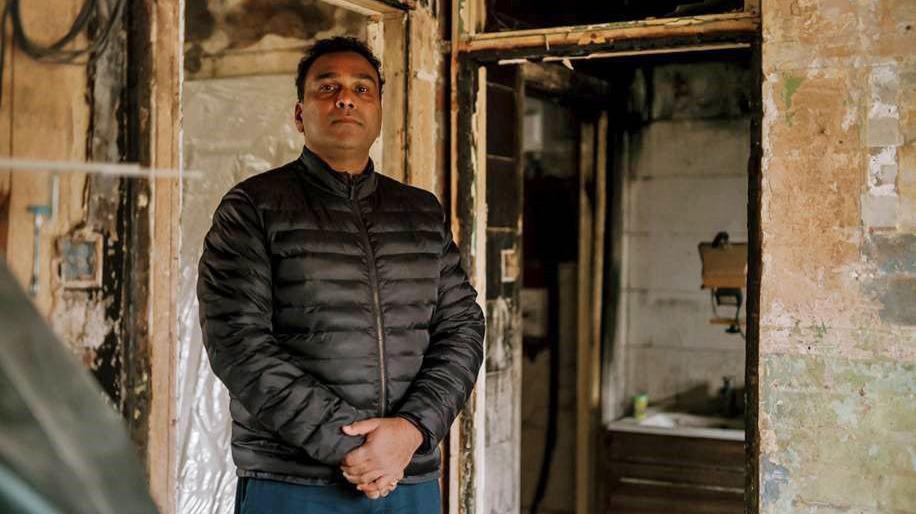 Don Hettiarachchi, a man with short dark hair wearing a dark padded jacket. He is standing inside his burnt-out home, which has charred door frames and walls and stripped-out electrics