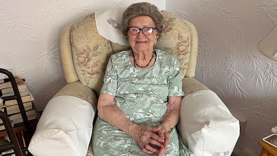 Averil Rowe. A woman with short brown hair sits in a chair. She is wearing glasses and is smiling