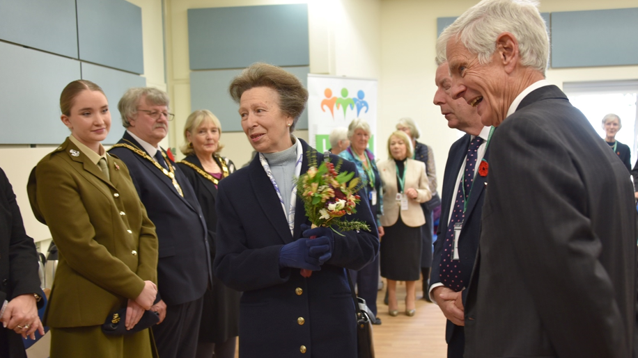 Princess Anne talks to people that are standing to welcome her in a large hall. She is carrying flowers.