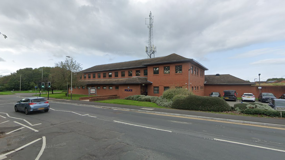 A two storey police station building with a car park to the right in front of a road