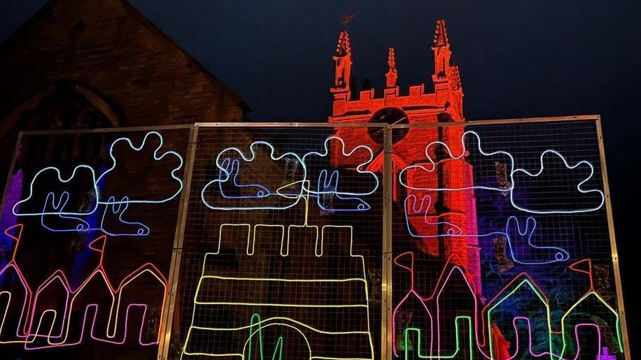 Illuminated artwork in front of a church featuring clouds and a yellow-coloured sandcastle 